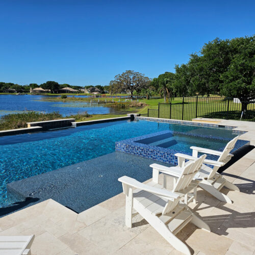 geometric pool with integrated spa. Two white outdoor lounge chairs face the pool with view of nearby lake
