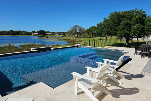 geometric pool with integrated spa. Two white outdoor lounge chairs face the pool with view of nearby lake