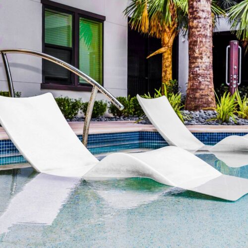 white pool lounge chairs submerged in shallow pool level with palm trees behind the pool